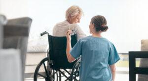 Pflegekraft spricht zu älteren Patientin, die im Rollstuhl sitzt. 