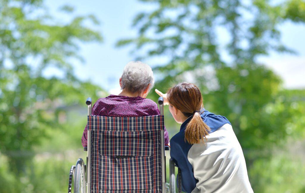 Eine Betreuungsassistentin zeigt einem älteren Menschen im Rollstuhl etwas in der Ferne. Beide sind von hinten zu sehen, umgeben von grünen Bäumen in einer friedlichen Umgebung.