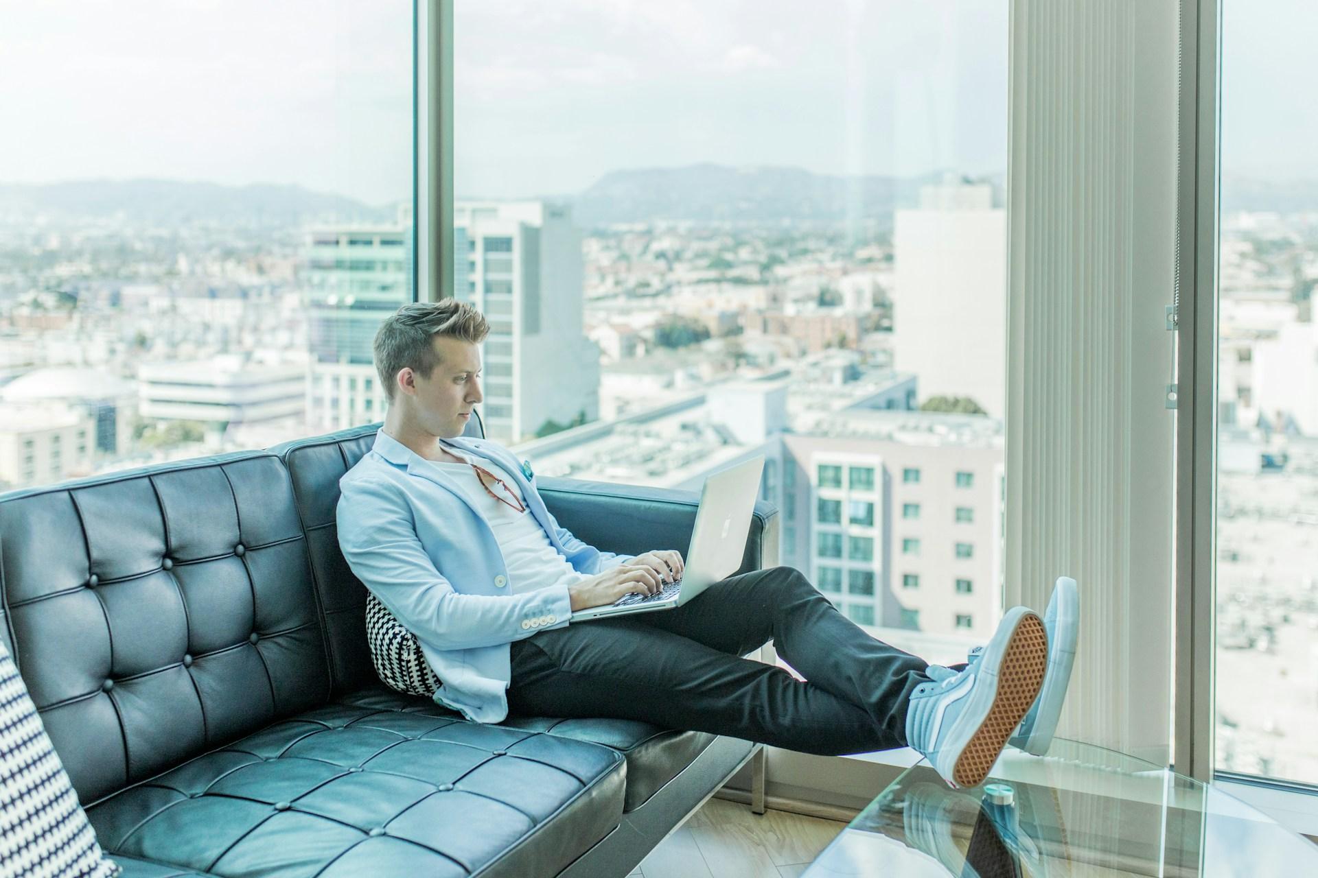 Ein Mann sitzt entspannt auf einem Sofa in einer modernen, lichtdurchfluteten Wohnung und arbeitet konzentriert an einem Laptop. Das Bild symbolisiert die Verbindung von Produktivität und Komfort in der heutigen Arbeitswelt, in der Flexibilität und technologische Unterstützung eine zentrale Rolle spielen.