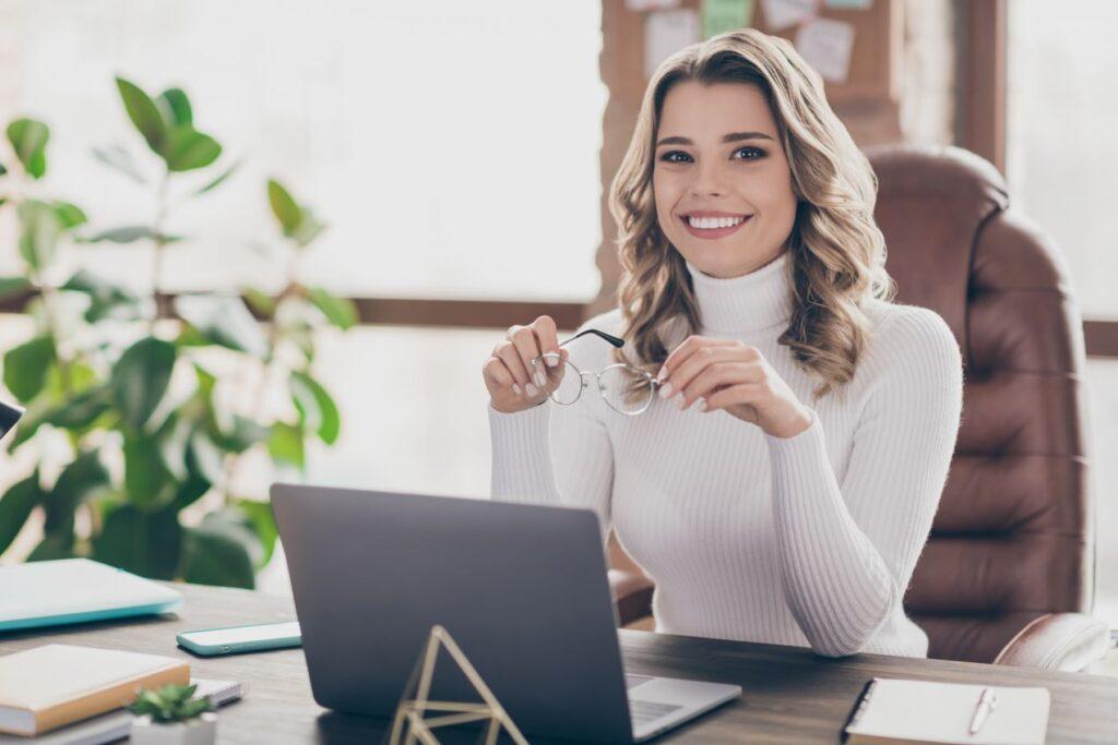 Lächelnde Frau am Schreibtisch mit Laptop und Brille, die für Compliance E-Learning-Lösungen arbeitet