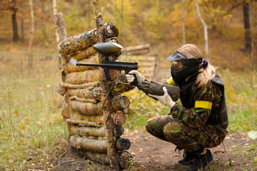 Spieler in Tarnkleidung zielt hinter Deckung mit Magfed Paintball Markierer im herbstlichen Gelaende, Fokus auf Ausruestung und Praezision