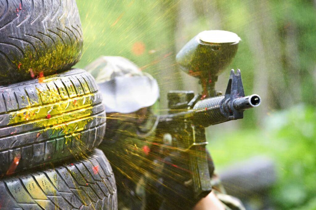 emf100 paintball spieler feuert auf gegner waehrend farbkugeln an reifen aufplatzen im gelände